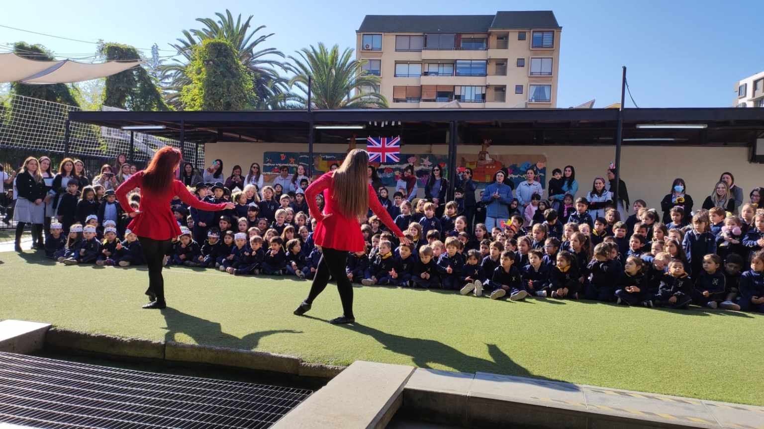Niños del Colegio TEI observando una presentación de baile en el patio, con bandera británica al fondo