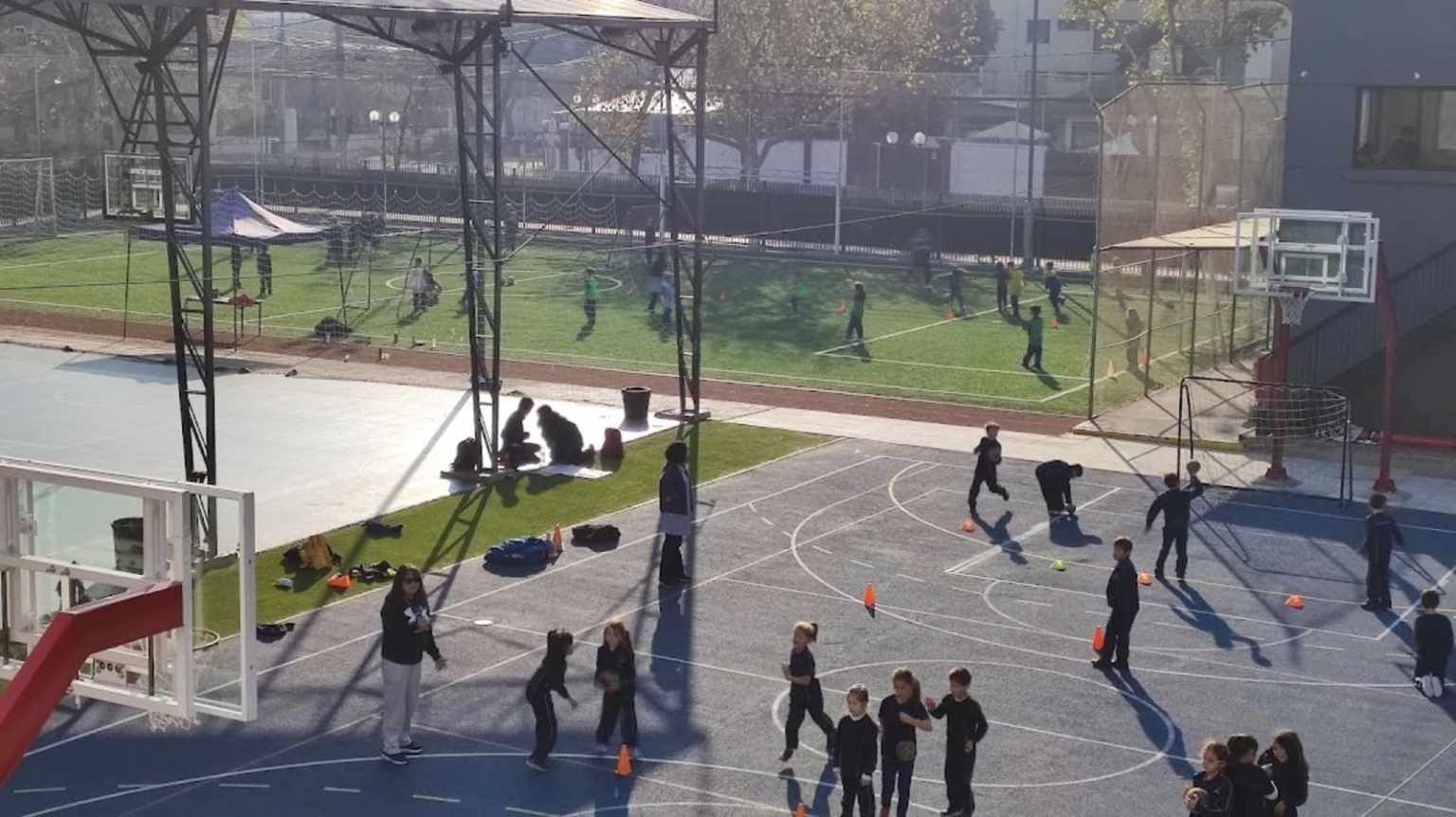 Estudiantes del Colegio TEI en clase de educación física en la multicancha, con conos y balones, y cancha de fútbol al fondo